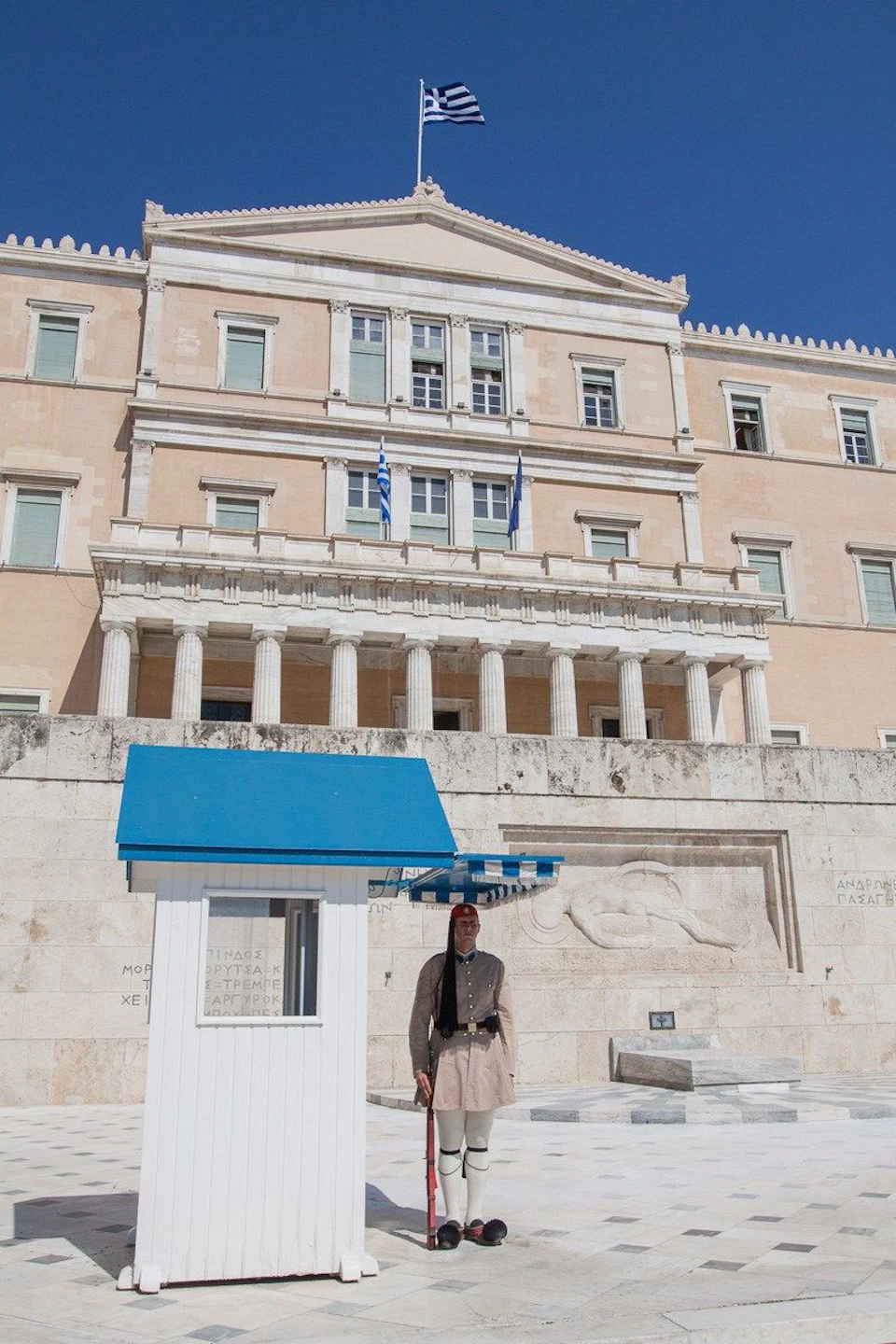 A guard stands by a small blue-roofed shelter in front of a neoclassical building with Greek flags and columns under a clear blue sky.