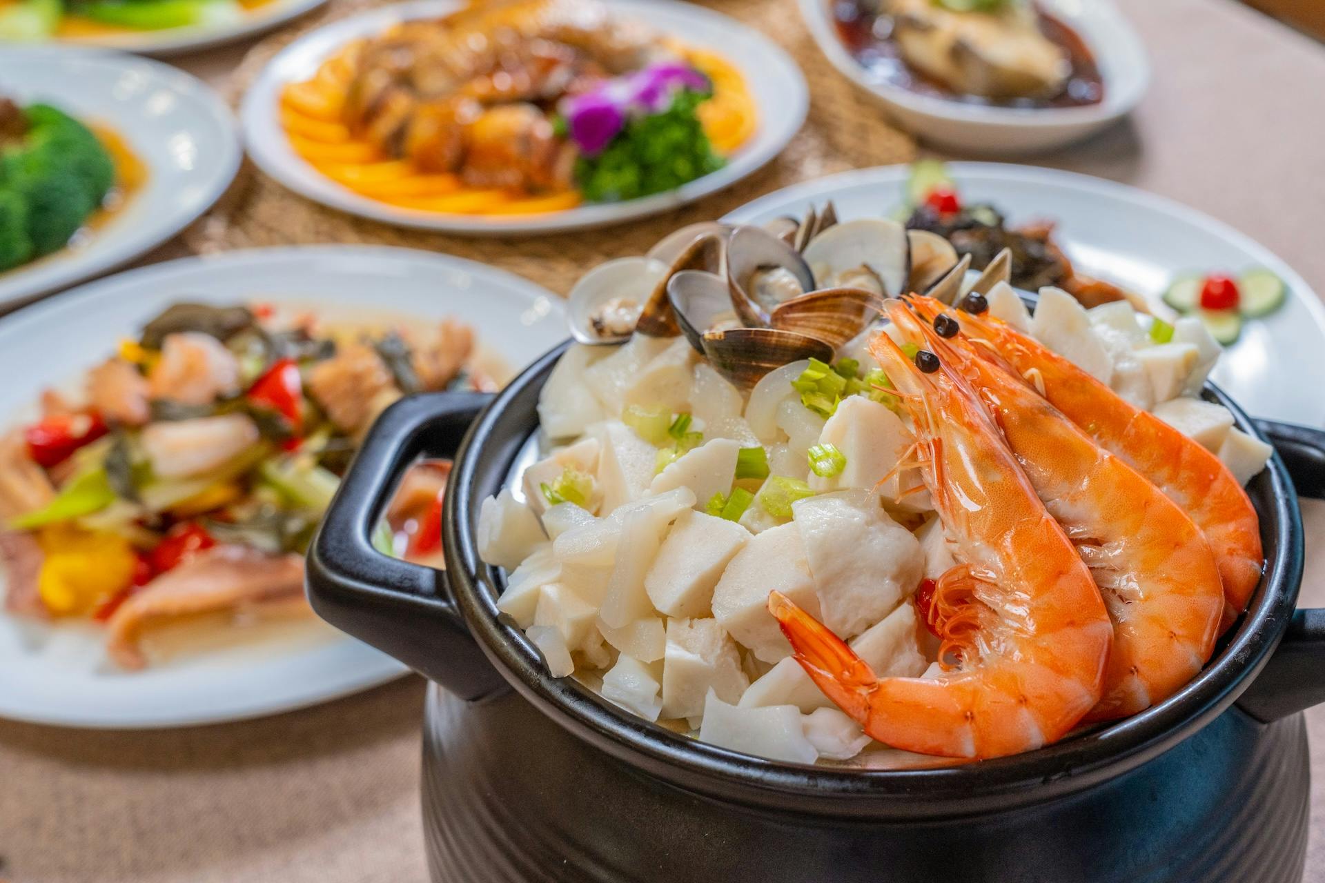 A seafood dish with shrimp, clams, and diced fish in a black pot, with other plated dishes blurred in the background.