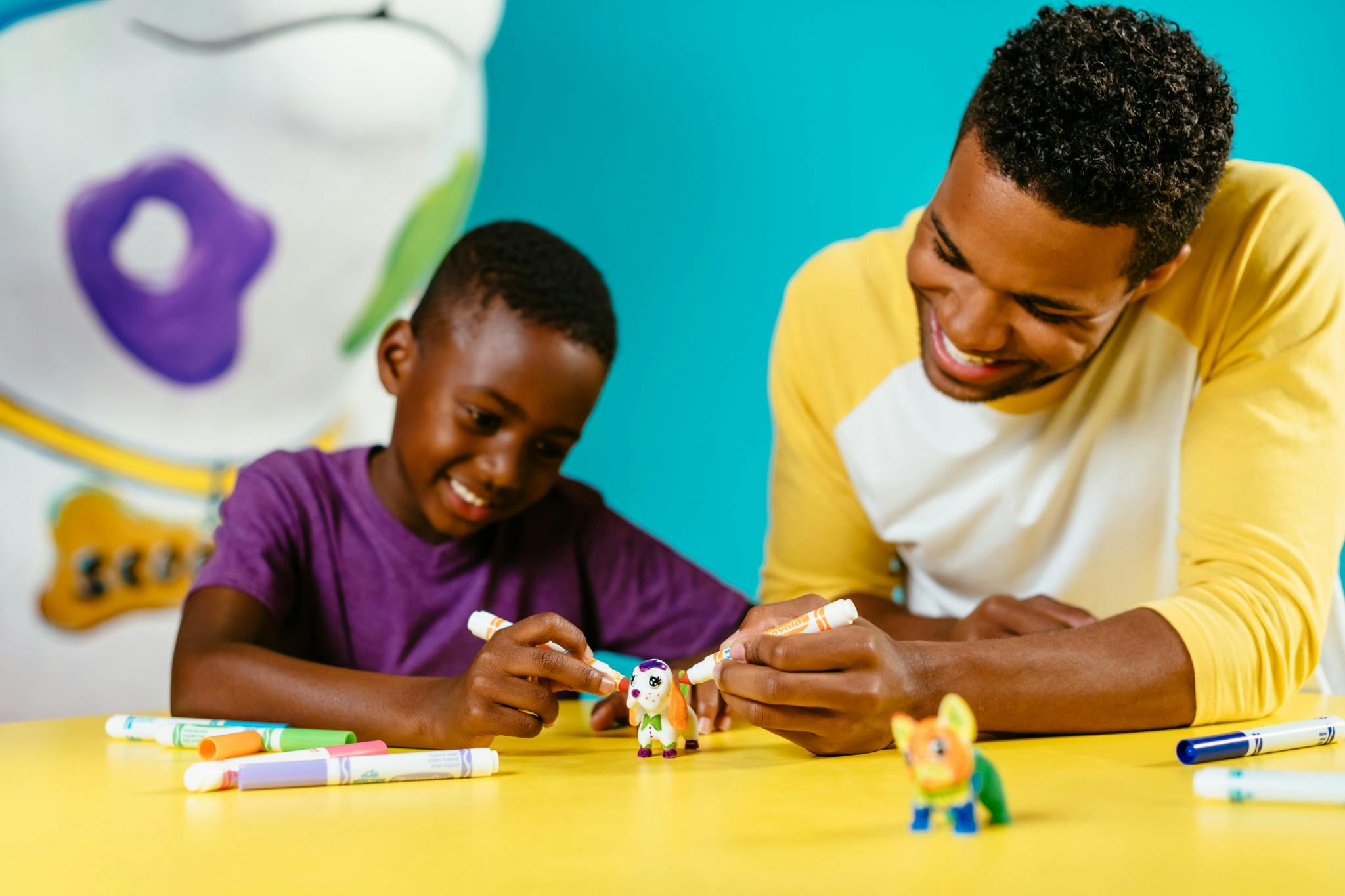 Two people smiling while coloring small toy figures with markers at a yellow table against a turquoise background.