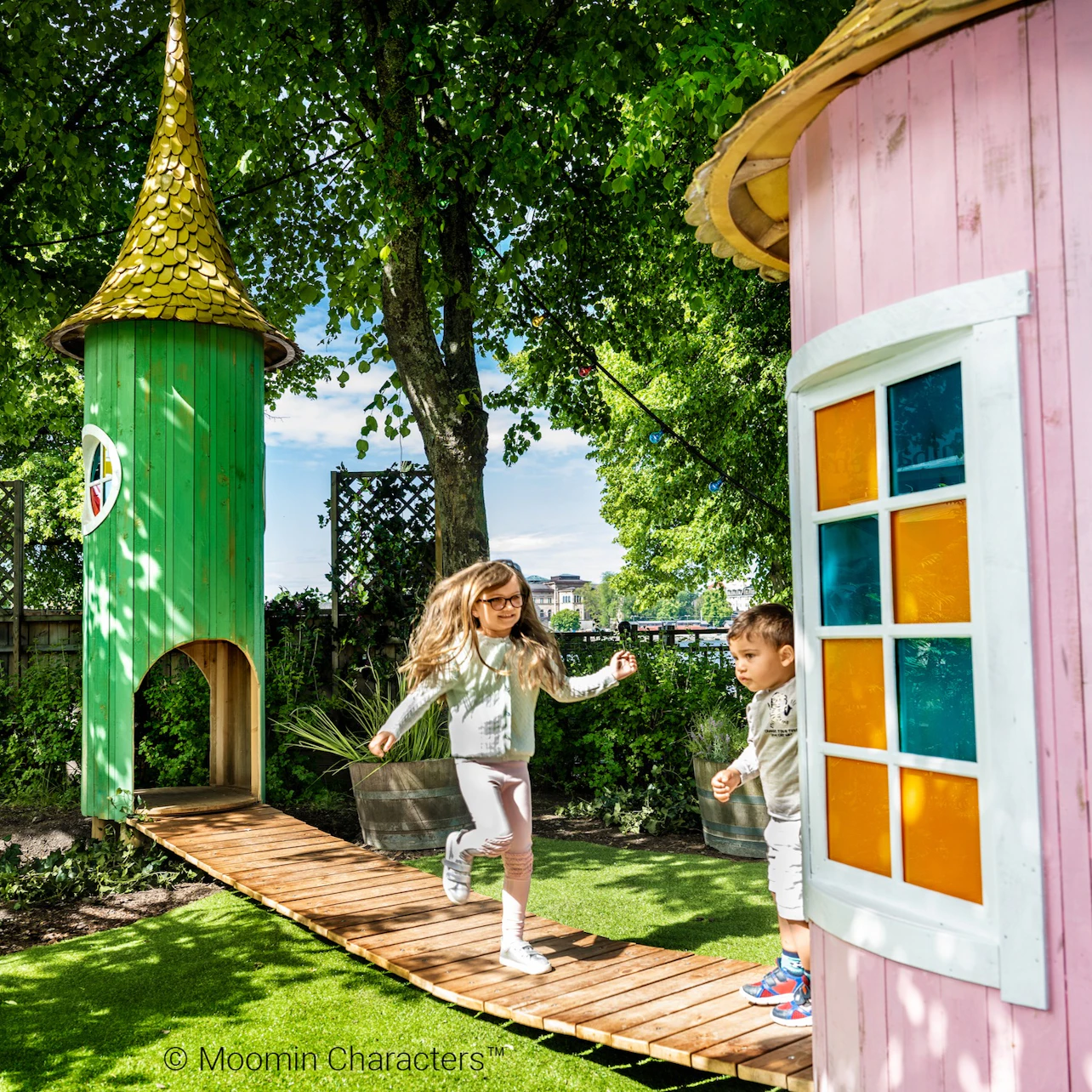 Two girls in white dresses interact between small wooden houses, one handing bread to the other. Tree and greenery in the background.