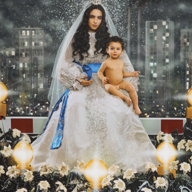 Uma mulher com um vestido branco ornamentado com uma faixa azul segura uma criança pequena, tendo como pano de fundo uma paisagem urbana e rodeada de flores e luzes.
