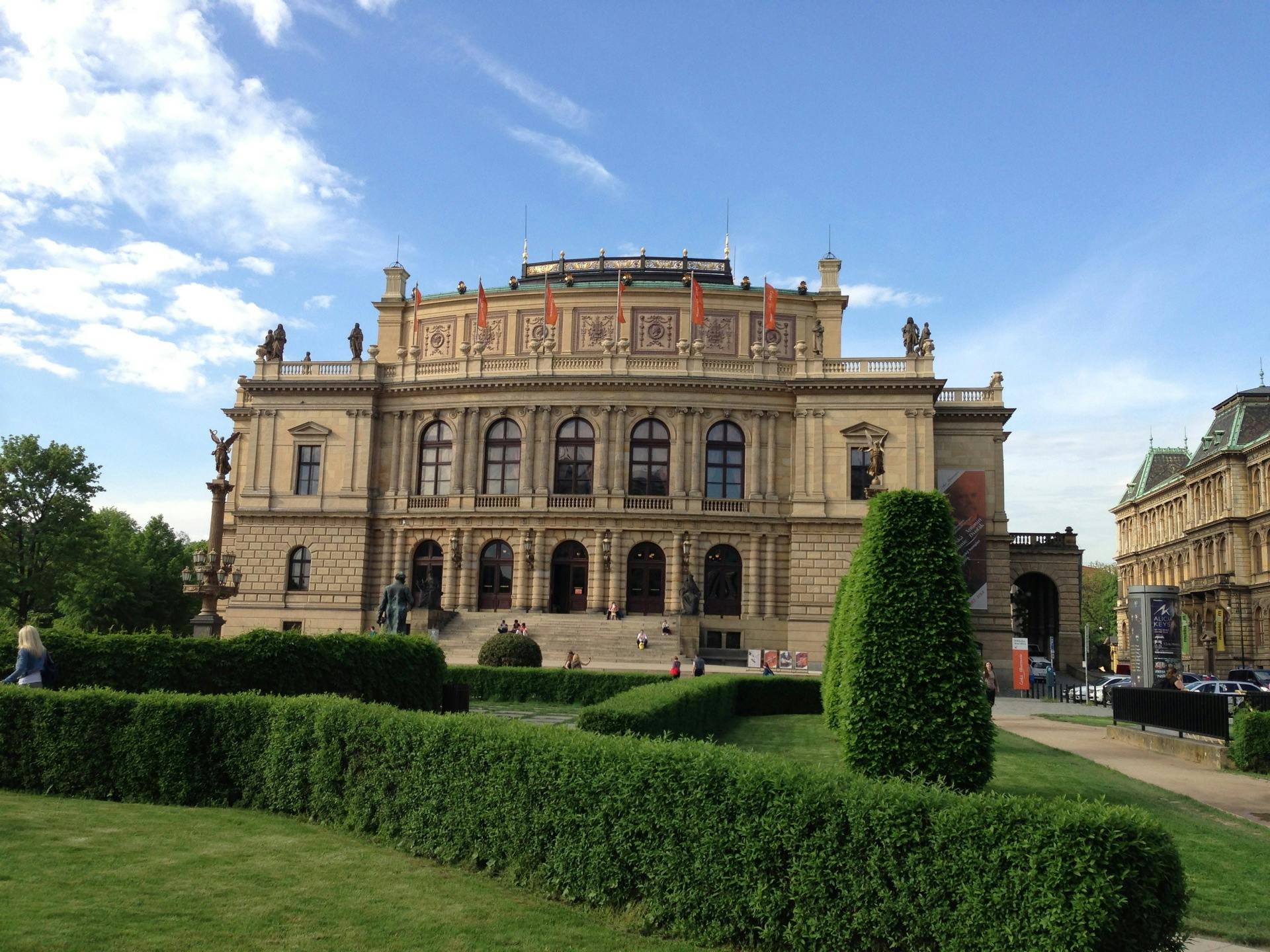 Large historic building with ornate design, green manicured bushes in front, people walking, and a clear blue sky.