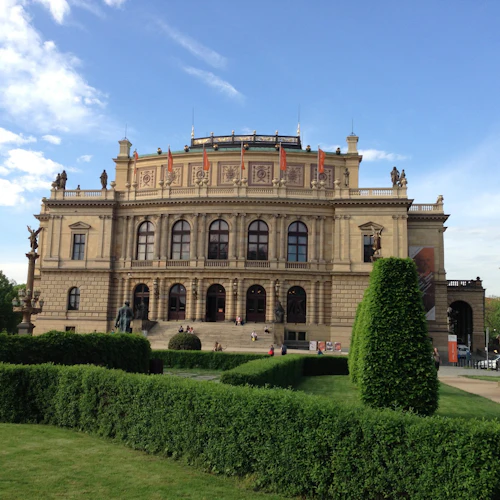Stor historisk byggnad med utsmyckad design, gröna välskötta buskar framför, människor som går och en klarblå himmel.