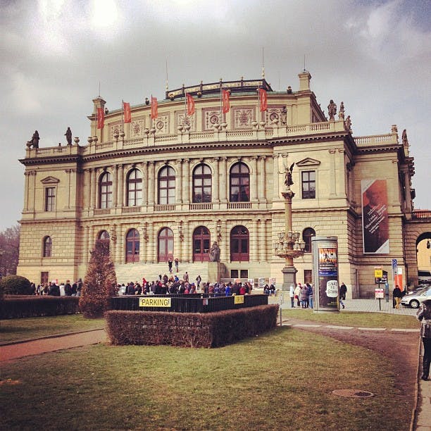 En historisk byggnad med välvda fönster, människor samlade utanför, en staty och flera skyltar, inklusive en för parkering.