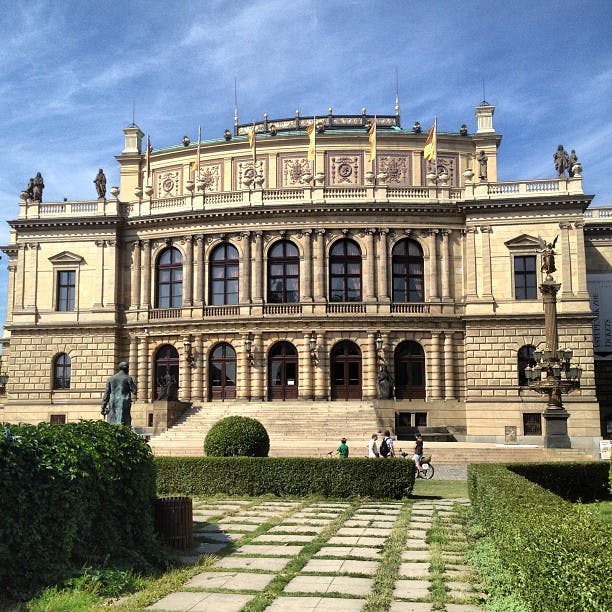 En stor, utsmyckad byggnad med pelare och statyer, frontad av en gångväg och buskar. Några personer promenerar och cyklar i närheten.