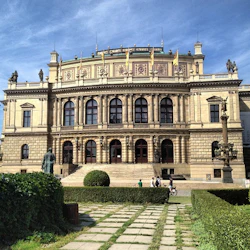 En stor, utsmyckad byggnad med pelare och statyer, frontad av en gångväg och buskar. Några personer promenerar och cyklar i närheten.