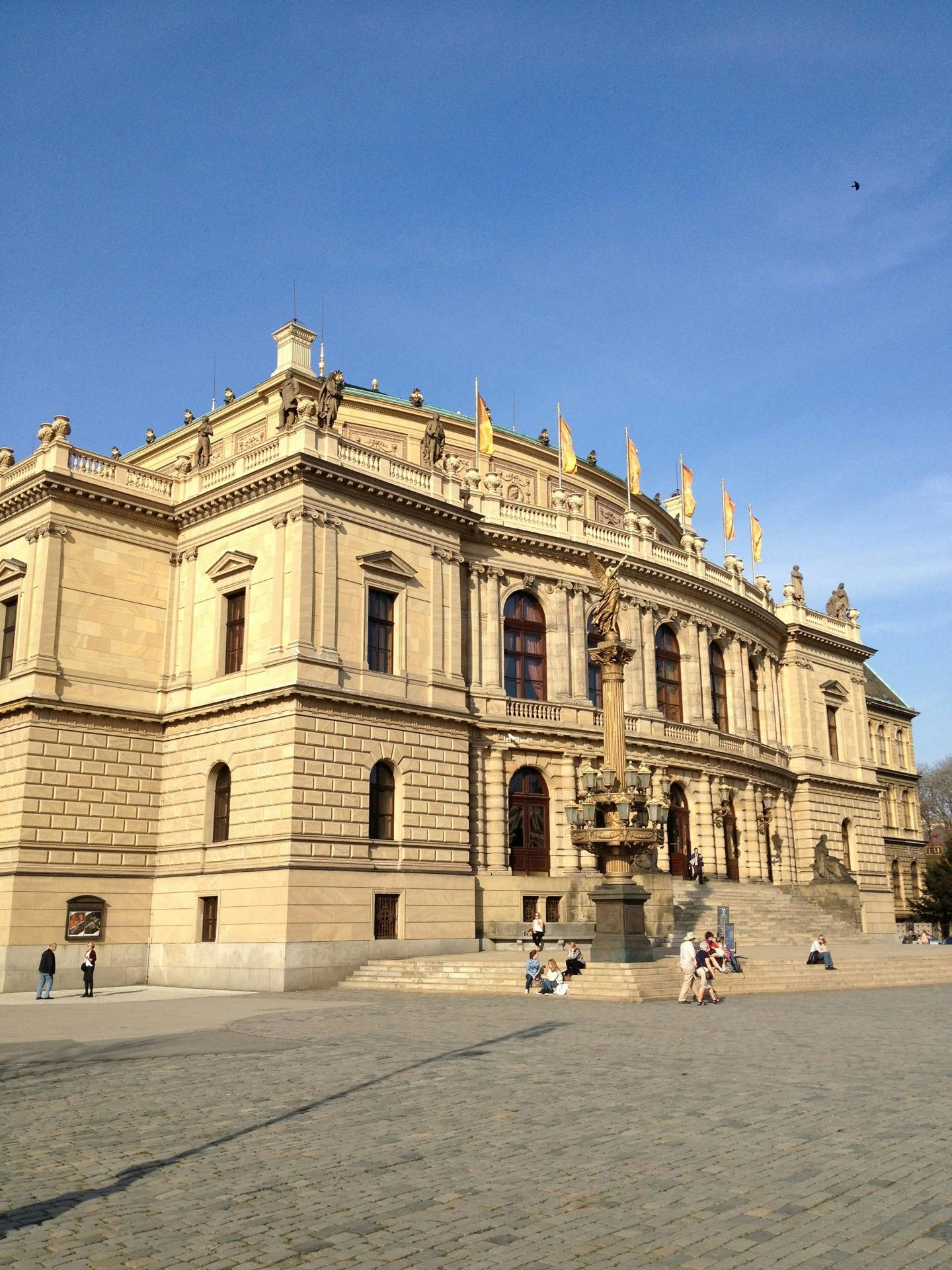 Stor historisk byggnad med utsmyckad arkitektur, gula flaggor på taket och människor som går och sitter nära entrétrappan.