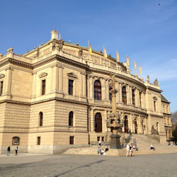 Stor historisk byggnad med utsmyckad arkitektur, gula flaggor på taket och människor som går och sitter nära entrétrappan.