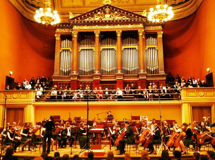 En symfoniorkester uppträder på scenen under en stor piporgel och ljuskronor, medan en publik tittar på från en balkong.