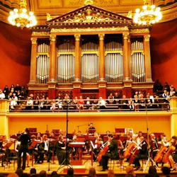 En symfoniorkester uppträder på scenen under en stor piporgel och ljuskronor, medan en publik tittar på från en balkong.