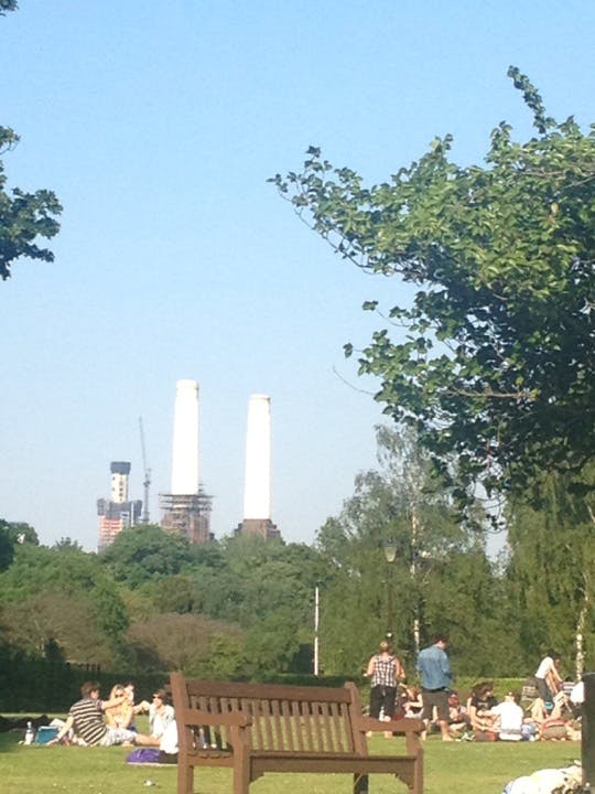 Människor som sitter och ligger på gräset i en park med två höga skorstenar och konstruktion i bakgrunden under en klar himmel.
