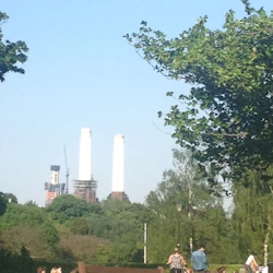 Människor som sitter och ligger på gräset i en park med två höga skorstenar och konstruktion i bakgrunden under en klar himmel.