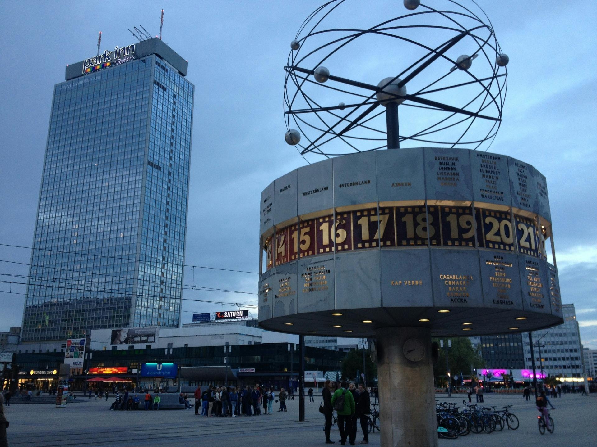 Hög glasbyggnad märkt "Park Inn" bredvid en stor världsklockstruktur med människor samlade på ett offentligt torg.