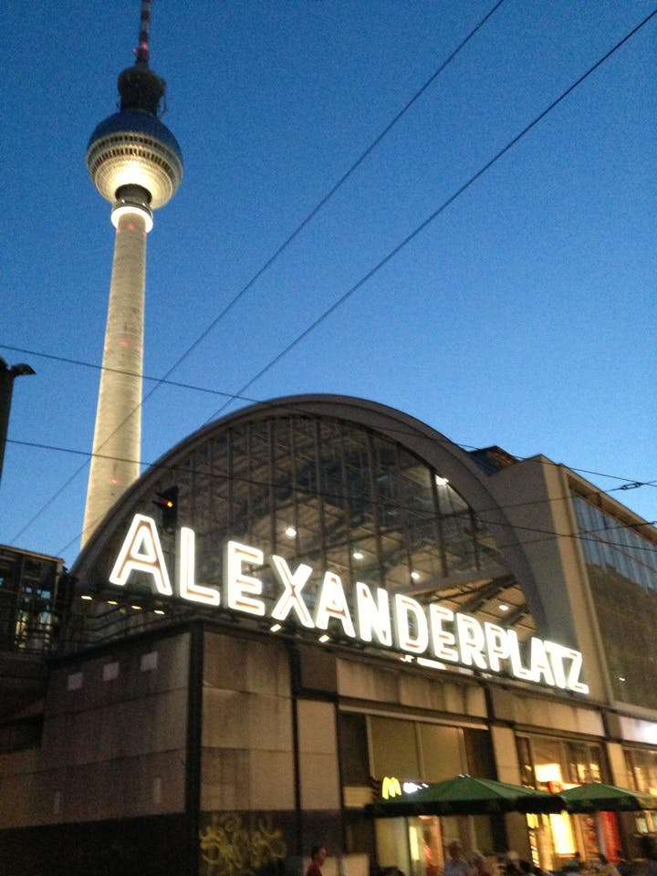 Alexanderplatz station lyste upp i skymningen med TV-tornet i bakgrunden mot en klarblå himmel.