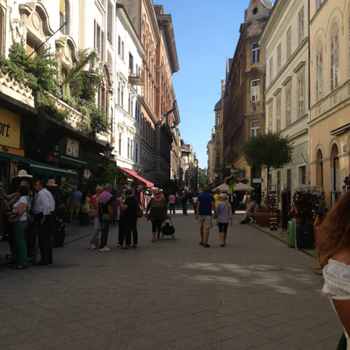 Gågata med människor som går, flankerad av historiska byggnader. Butiker och grönska pryder vänster sida, under en klarblå himmel.