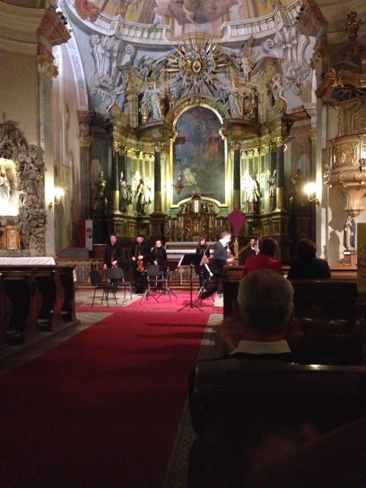 Orkestermedlemmar står framför ett utsmyckat kyrkoaltare, med en publik sittande på träbänkar.