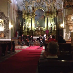 Orkestermedlemmar står framför ett utsmyckat kyrkoaltare, med en publik sittande på träbänkar.