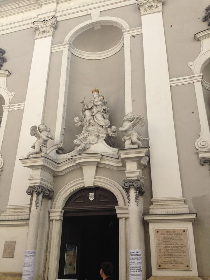 Utsmyckad kyrkoentré med statyer av änglar och en central figur ovanför dörren. En person går in i kyrkan.