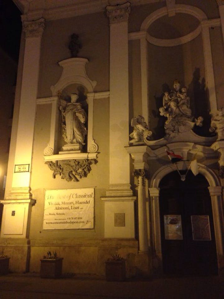 Upplyst stenfasad med klassiska statyer, en konsertbanner och en mörk utsmyckad dörröppning med flaggor på natten.