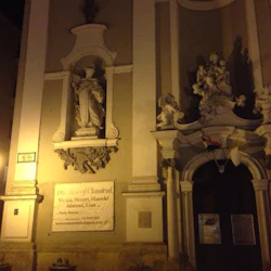 Upplyst stenfasad med klassiska statyer, en konsertbanner och en mörk utsmyckad dörröppning med flaggor på natten.