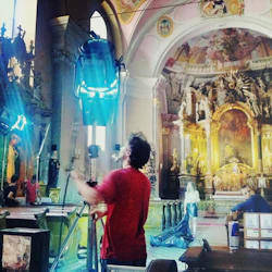 En man justerar belysningsutrustning inuti en utsmyckad kyrka, med detaljerade religiösa statyer och ett stort altare i bakgrunden.