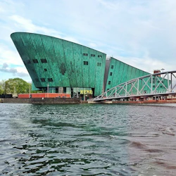 En stor grön, båtformad byggnad märkt "NEMO" med en gångbro över vatten under en delvis molnig himmel.