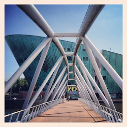 Människor som går på en bro med en geometrisk ram som leder till en modern, grön, båtformad byggnad under en klarblå himmel.