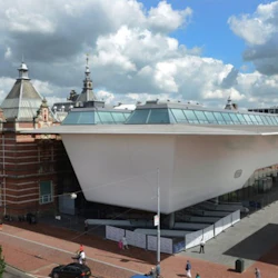 En modern vit byggnad med sluttande tak intill en historisk tegelbyggnad under en delvis molnig himmel. Folk går och cyklar nedanför.