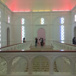 Människor som står och går i ett modernt, starkt upplyst galleri med färgglada neonljus och intrikata vita räcken.