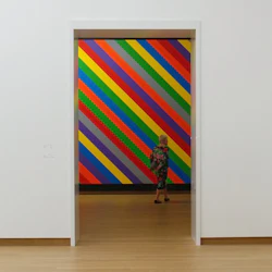 En person står framför en vägg med färgglada diagonala ränder i ett konstgalleri.