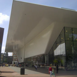 En modern byggnad med ett stort vitt överhäng och glasväggar. Folk går nedanför på den rymliga trottoaren.