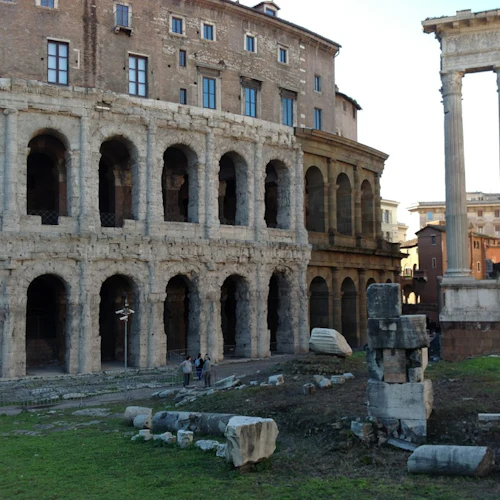 Forntida romerska ruiner med välvd struktur, stående kolonner och utspridda stenrester; några personer synliga i bakgrunden.