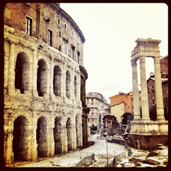 Forntida romerska ruiner med en flervalvad struktur och två stående pelare, ställda mot moderna byggnader.