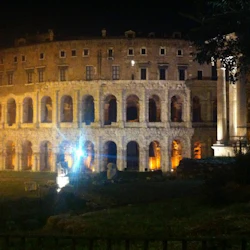 En antik romersk amfiteater upplyst på natten, med valv och en närliggande pelare, mot en mörk himmel.