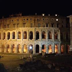 Forntida amfiteater upplyst på natten med rader av valv, en stor pelare och utspridda ruiner i förgrunden.