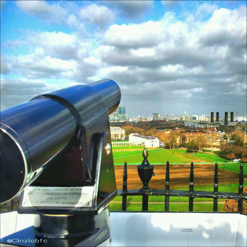 Czarny teleskop na balkonie z widokiem na park z drzewami i budynkami pod zachmurzonym niebem.