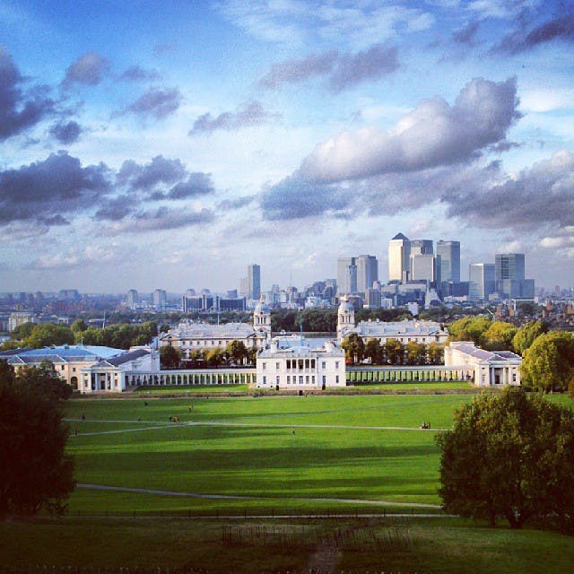 En historisk byggnad med vita pelare och torn, stora gröna gräsmattor, träd och avlägsna moderna skyskrapor under en delvis molnig himmel.