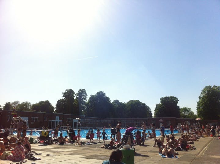 Crowded outdoor pool area with people swimming and sunbathing on a sunny day. Trees and a building in the background.