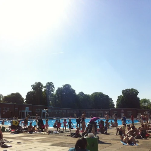 Trångt utomhuspoolområde med människor som badar och solar en solig dag. Träd och en byggnad i bakgrunden.