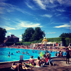 Fullsatt utomhuspool under en klarblå himmel. Folk badar, lounge, och sola. Livräddare övervakar från en hög stol.