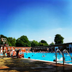 Människor kopplar av och badar i en utomhuspool under en klarblå himmel, omgiven av röda tegelbyggnader och gröna träd.