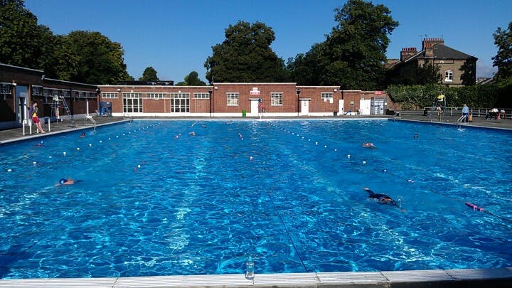 En utomhuspool med gränder, simmare och en tegelbyggnad i bakgrunden, omgiven av träd under en klar himmel.