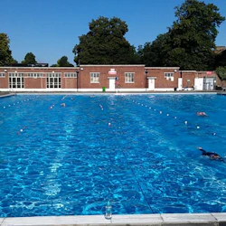 En utomhuspool med gränder, simmare och en tegelbyggnad i bakgrunden, omgiven av träd under en klar himmel.