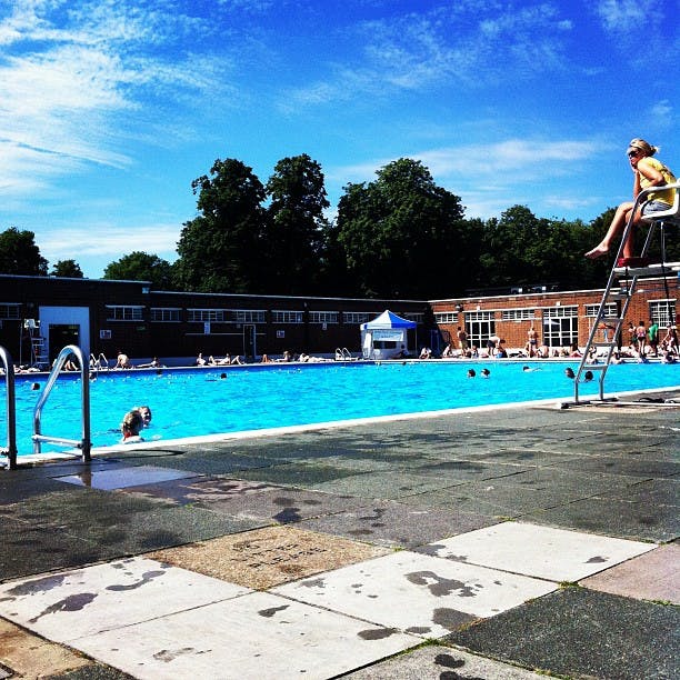 Utomhuspool med människor som simmar och solar. En badvakt sitter på en barnstol. Träd och byggnader i bakgrunden.