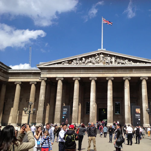 En grupp människor samlas framför en byggnad med pelare och en brittisk flagga ovanpå, under en blå himmel.