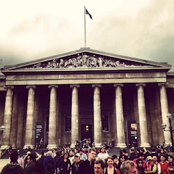 Trängsel framför en storslagen byggnad med höga pelare, dekorativ frise och en flaggstång en molnig dag.