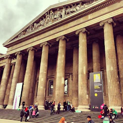 Människor går uppför trappor till en stor byggnad med grekiska kolumner och utsmyckade prydnadsföremål, med banderoller som främjar en utställning.