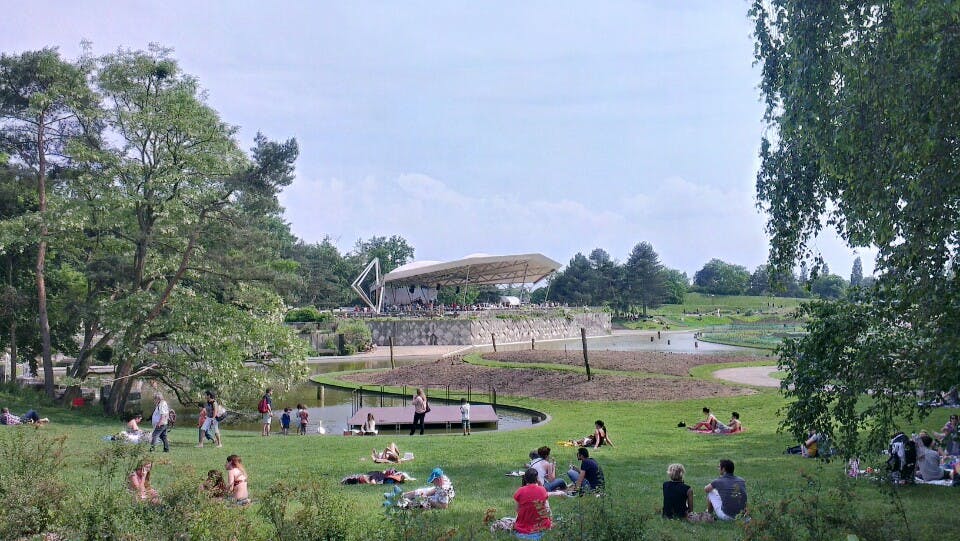 Des personnes se détendent sur une pelouse dans un parc avec des arbres, de l'eau et une scène accueillant un événement en arrière-plan.