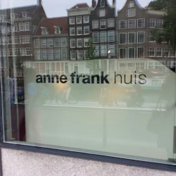 Bilden visar ett fönster med texten "Anne Frank Huis", som speglar kanalhus och människor på en gata i Amsterdam.
