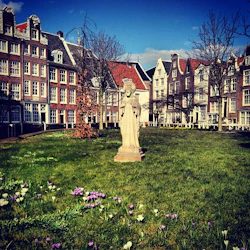 En stenstaty står på en gräsbevuxen gräsmatta prydd med lila och vita blommor, omgiven av historiska byggnader i europeisk stil.